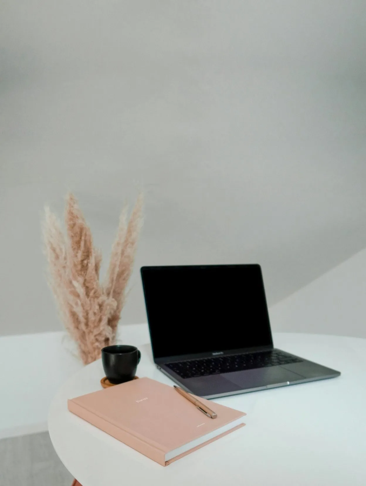 Laptop with a dark screen on a round white table beside pampas grass, a black cup and a pink notebook