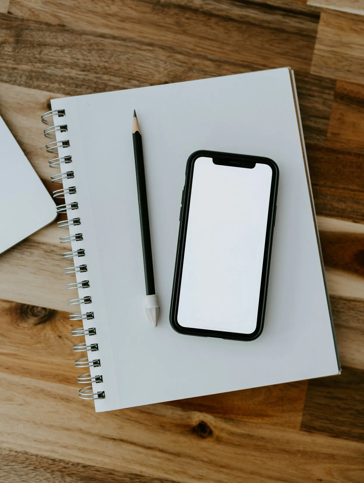 Smartphone with a blank white screen lying on a spiral notebook beside a pencil on a wooden desk