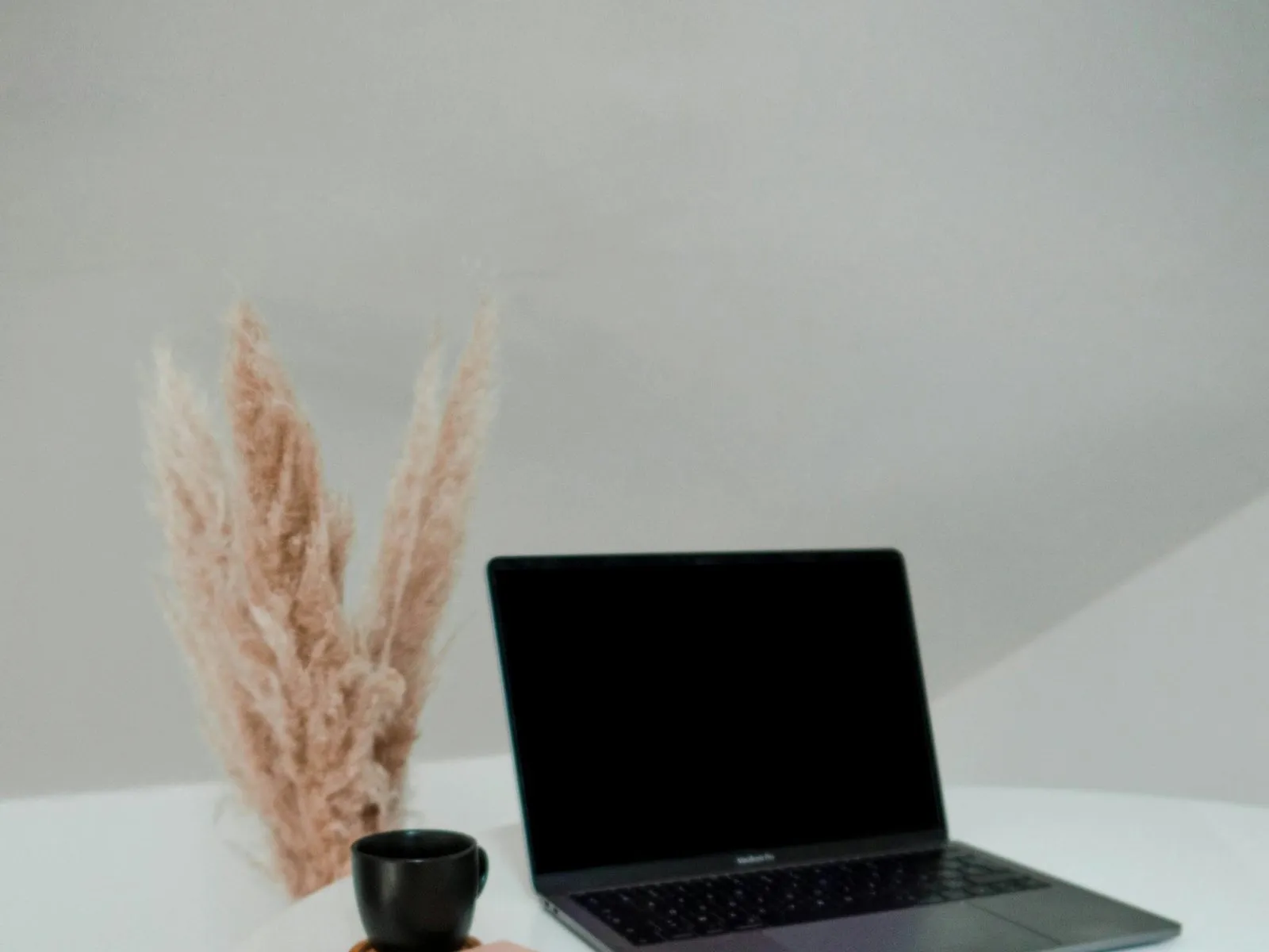 Laptop with a dark screen on a round white table beside pampas grass, a black cup and a pink notebook