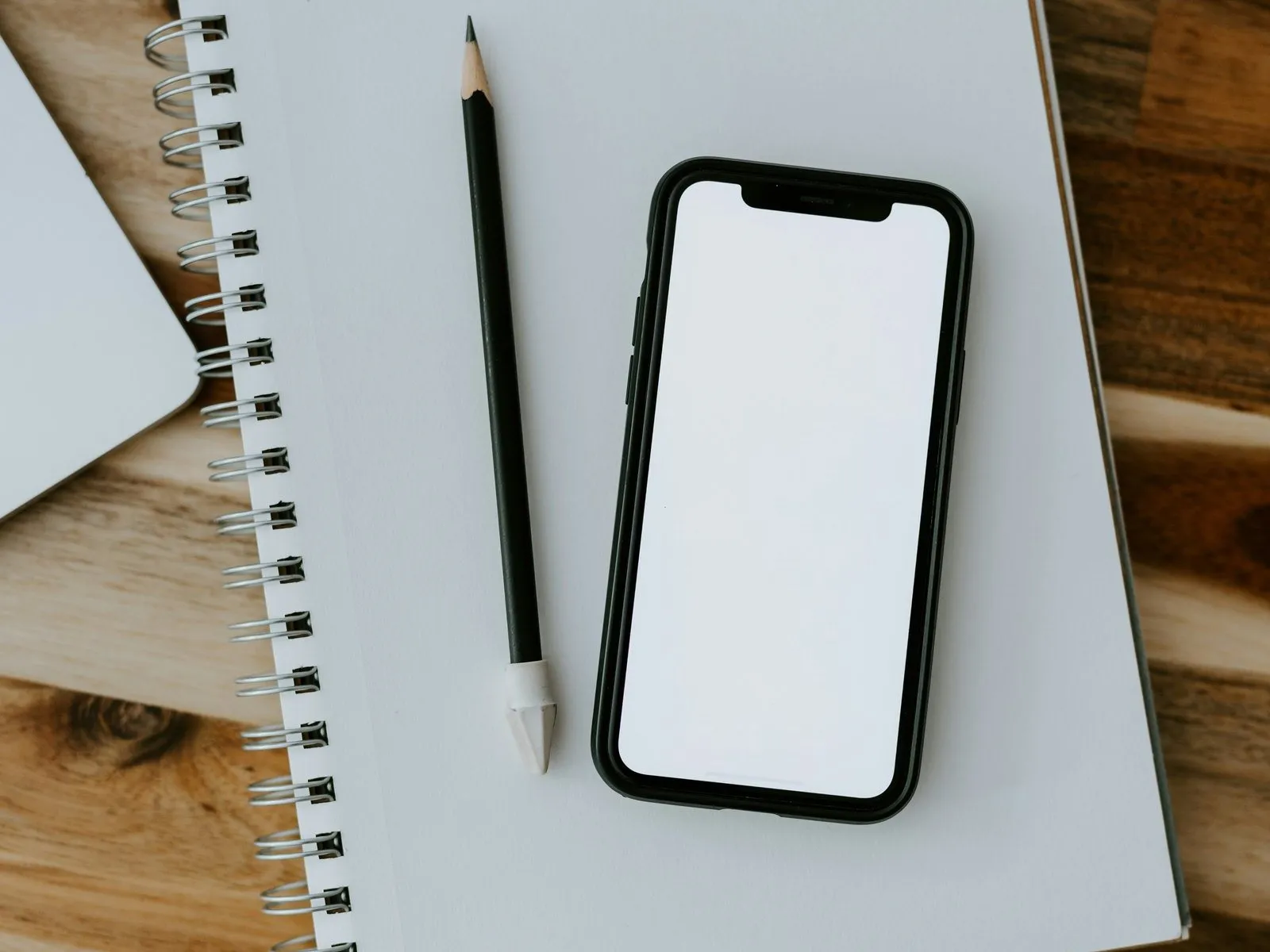 Smartphone with a blank white screen lying on a spiral notebook beside a pencil on a wooden desk